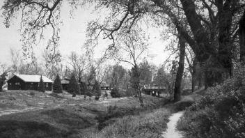 Black-and-white photograph of a country footpath by a stream, cottages, and large trees.