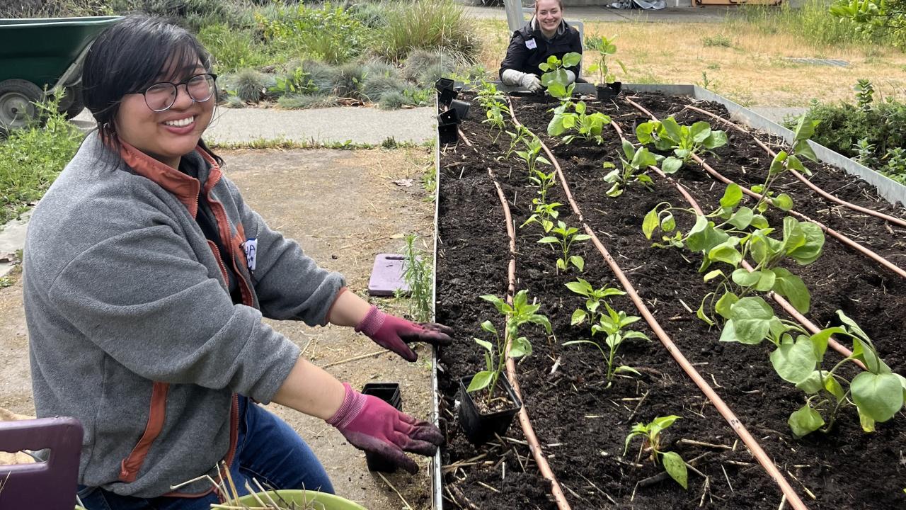 Two smiling people kneel next to a garden bed at the Good Life Garden
