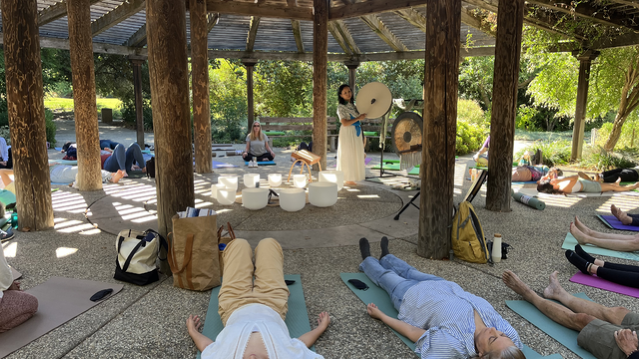 People lying on mats under wooden pergola in outdoor relaxation session with singing bowls