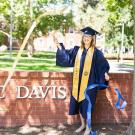 Image of UC Davis grad in cap, gown and stole waving blue and gold streamers in front of the brick UC Davis monument.