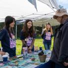 Three smiling people in purple shirts at outdoor outreach table talking to a visitor