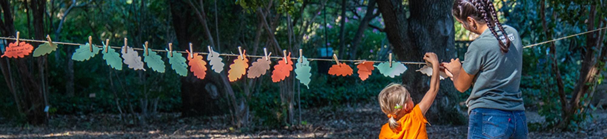 Image of student helping child place oak leaf on laundry line in UC Davis Shields Oak Grove.